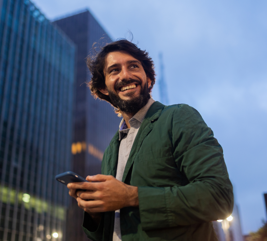 A young business man on a city street smiling and holding his phone while he checks on his business money market account through his mobile app