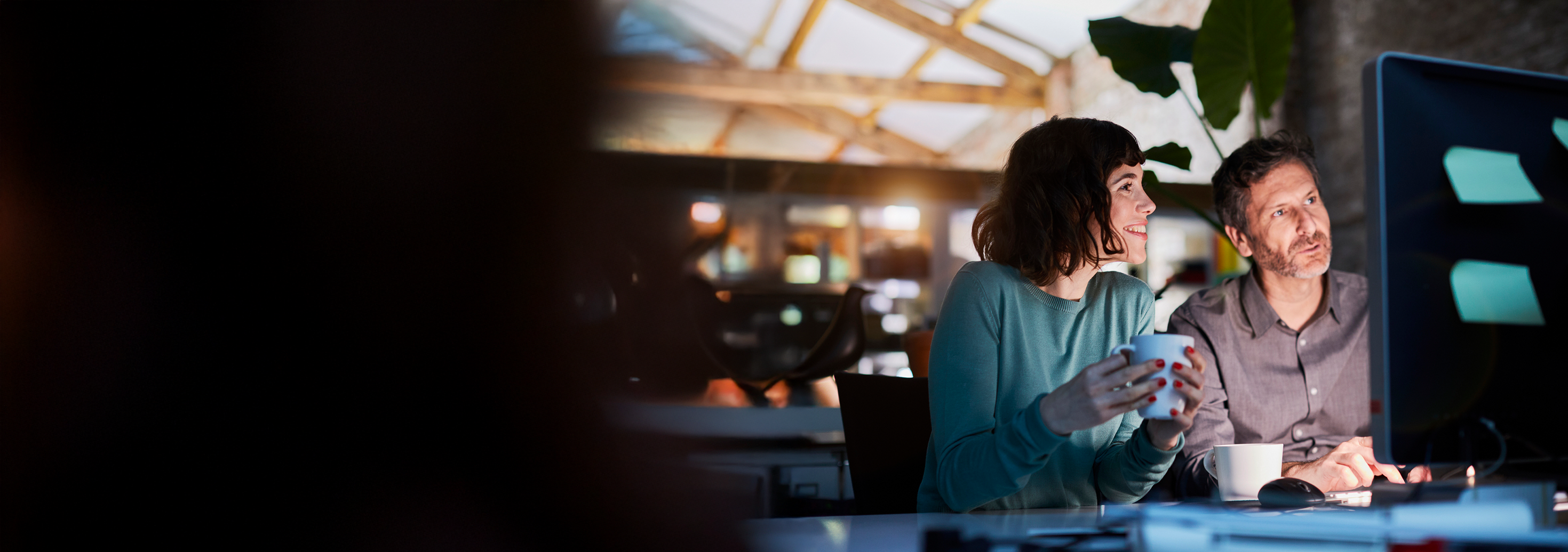 a man and a woman, at a desk drinking coffee and reading a computer monitor, perhaps weighing their investment solutions options for the future by reviewing our website