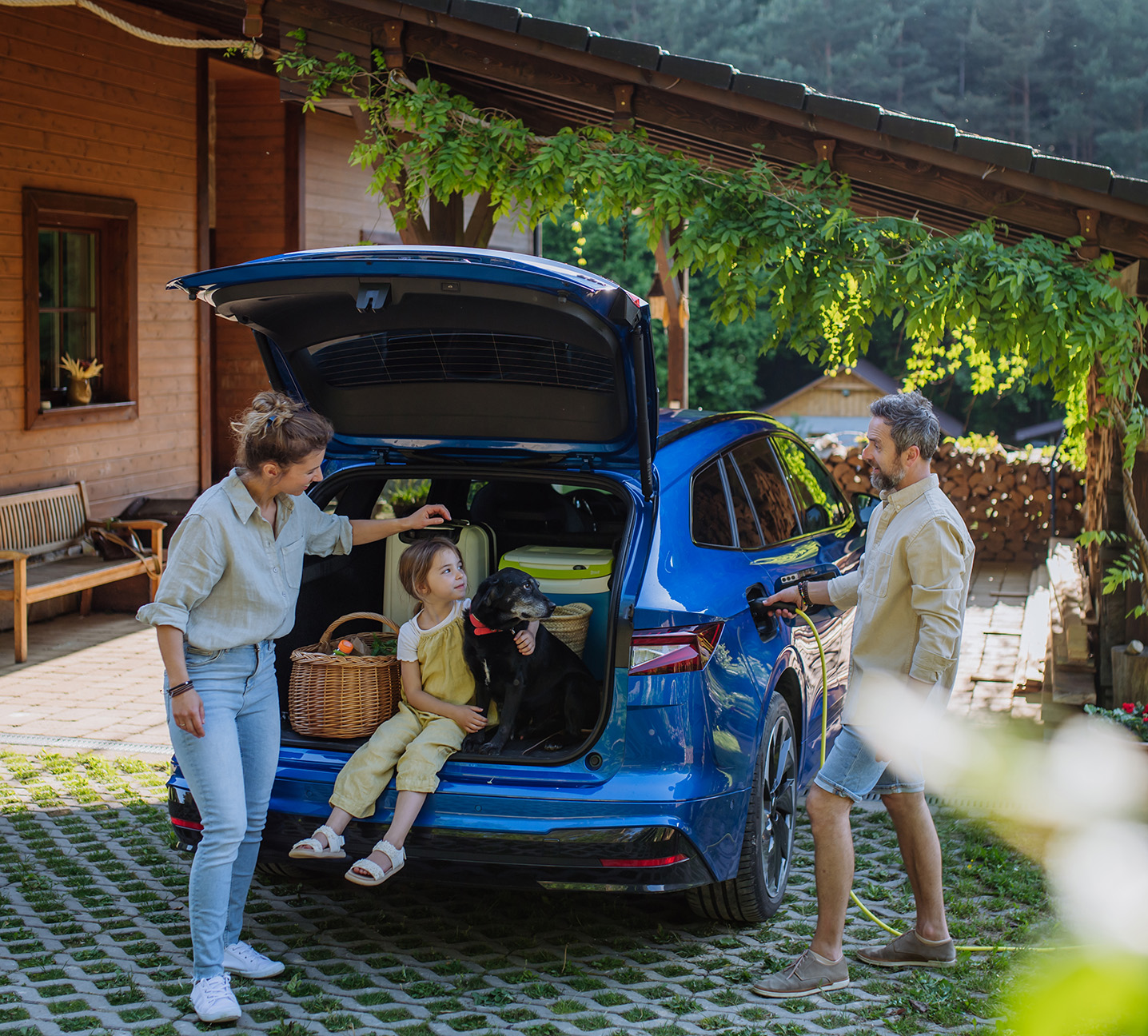 Father charging their electric car, little daughter and mother sitting and waiting in car trunk.