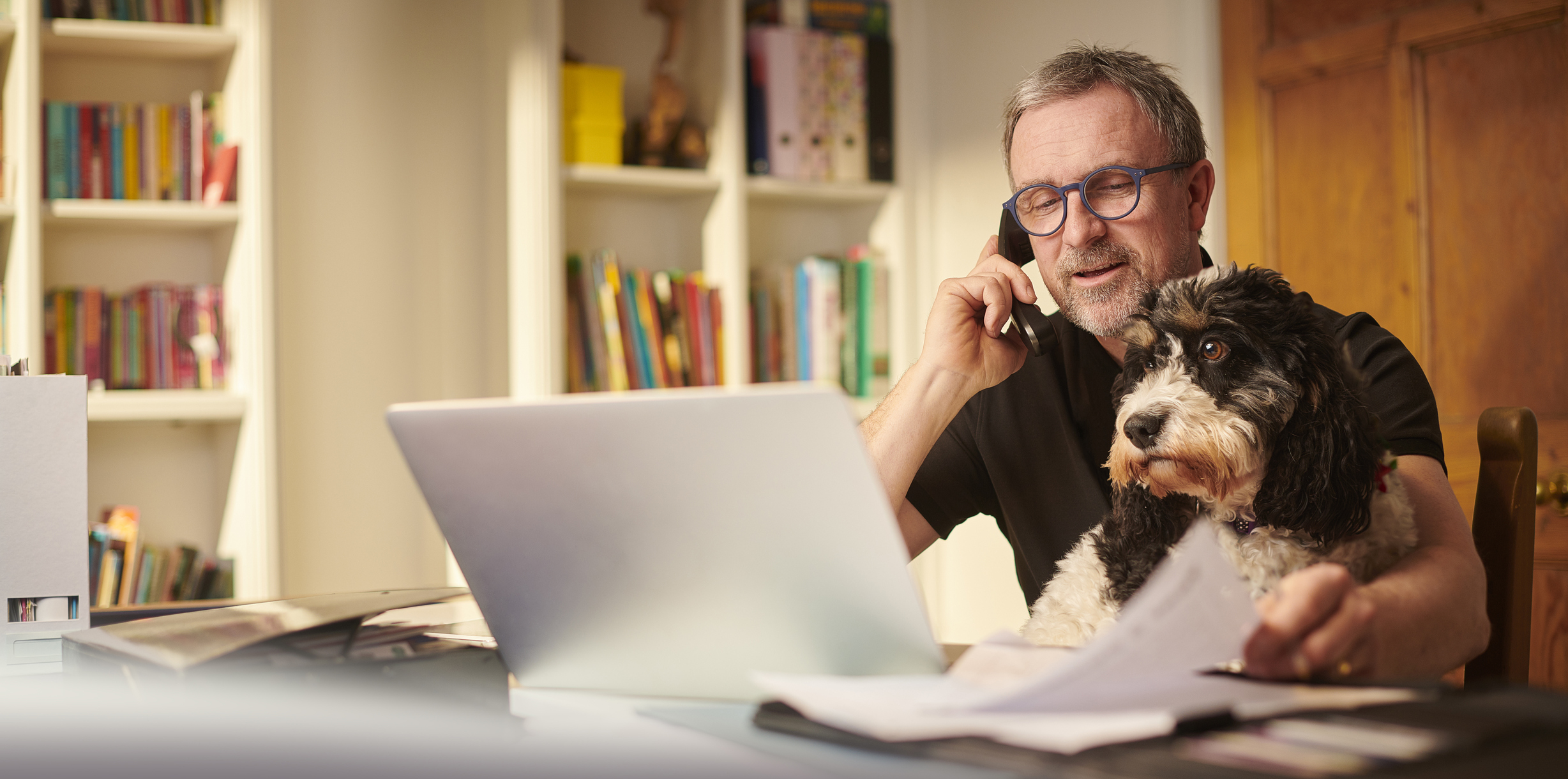 A man and his dog are sitting at his desk using his laptop while on the phone with PACU to ask us how to switch his accounts to Piedmont Advantage.